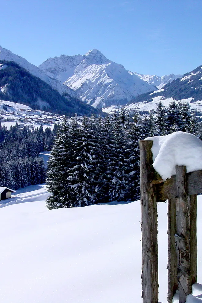 Easter Offer Snow-covered alpine landscape with wooden fence, trees, cabin, and mountain village in the background